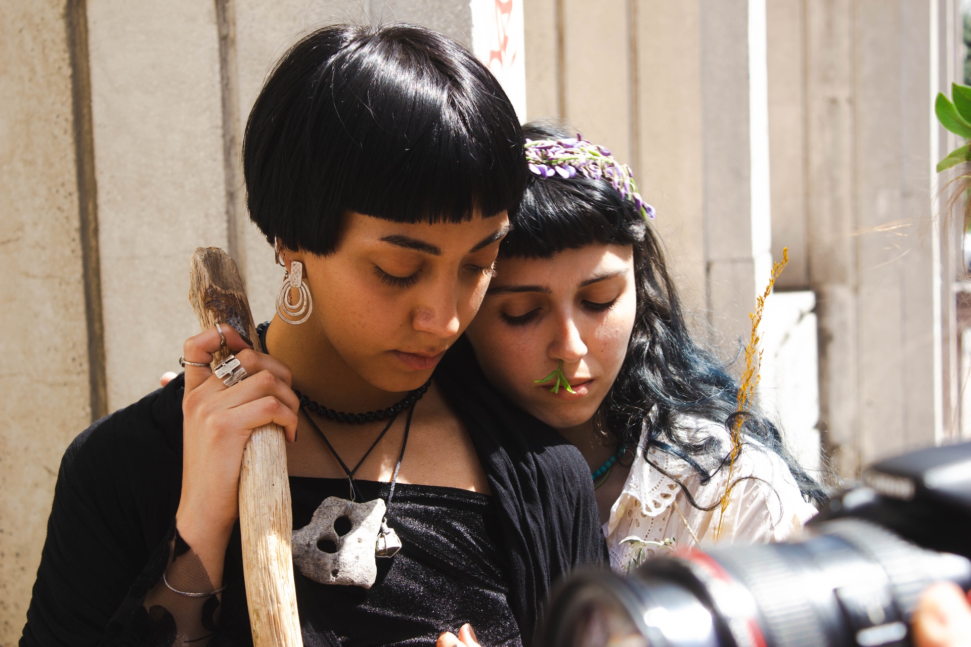 Enacting Ourselves — two performers lean close together in a sunlit outdoor courtyard, one holding a wooden staff and wearing a stone mask pendant