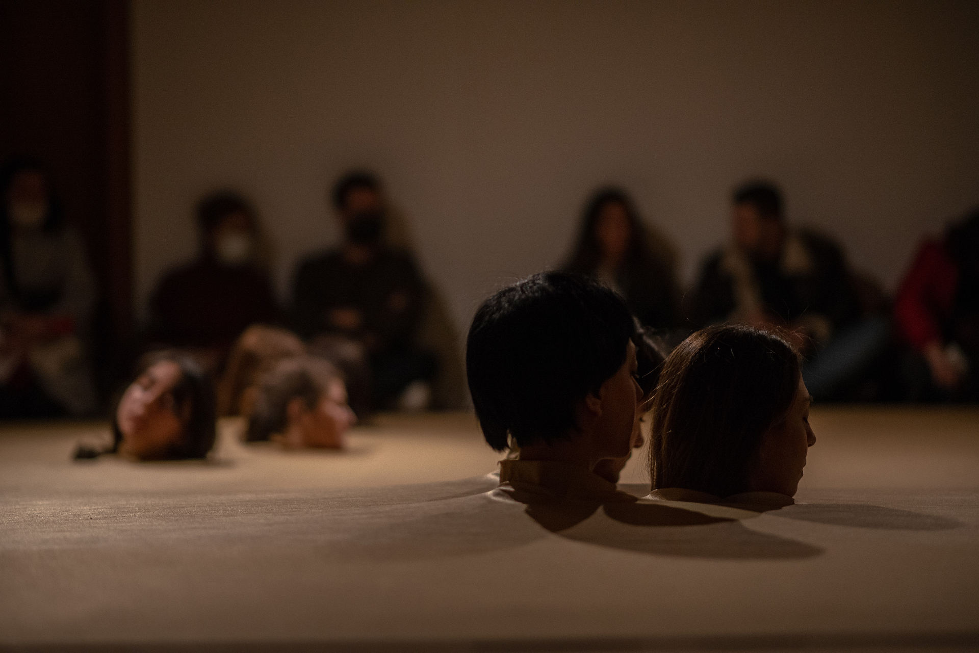 Head to Head — performers lying on the floor with heads emerging from paper while an audience watches in a dim gallery